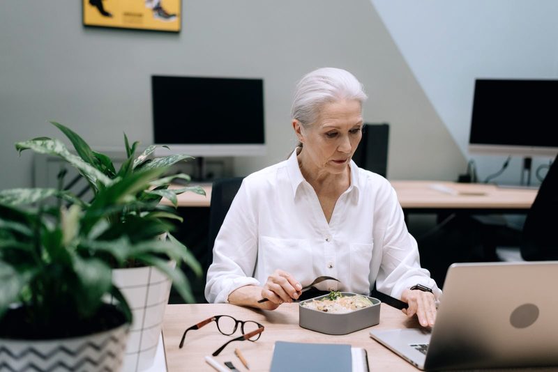 Frau mit weißem Haar beim Mittagessen an ihrem Schreibtisch