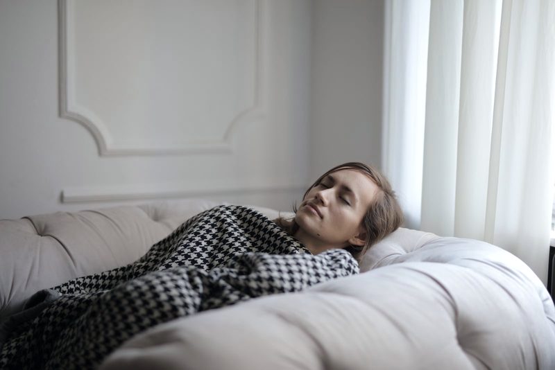 Frau liegt am Abend mit geschlossenen Augen unter einer Decke auf dem Sofa